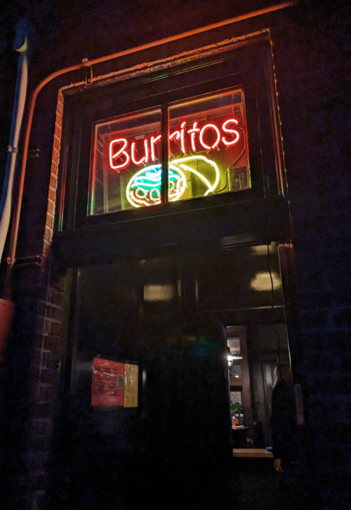 A brightly lit neon sign reading "Burritos" in a window at night, with a dark brick wall in the background.