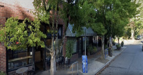 A street view of a brick building with large windows, striped awnings, and trees lining the sidewalk.