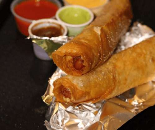 Two crispy, golden-brown spring rolls on foil, accompanied by small cups of various dipping sauces.