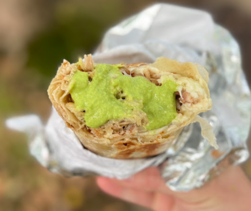 A hand holding a partially unwrapped burrito filled with rice, meat, and green sauce.