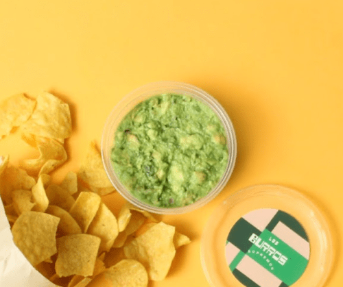 A container of guacamole next to a pile of tortilla chips on a bright yellow background.