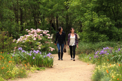 Two women walk along a garden path surrounded by colorful flowers and lush greenery.