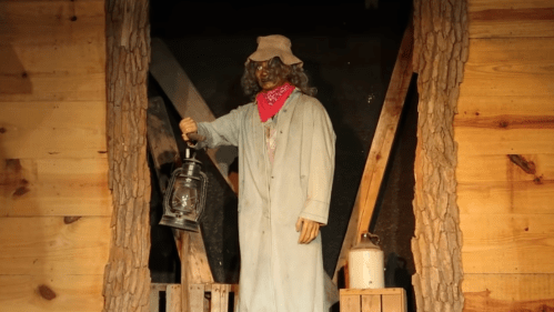 A life-sized figure in a blue coat and hat holds a lantern, standing in a rustic wooden setting.