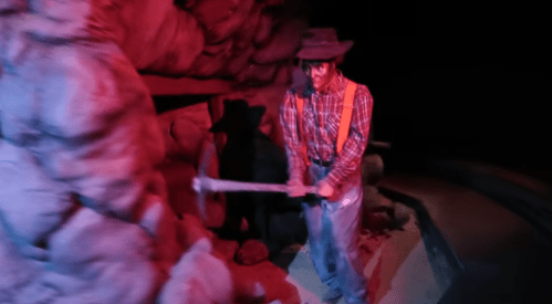 A miner in a plaid shirt and hat swings a pickaxe in a dimly lit cave setting.