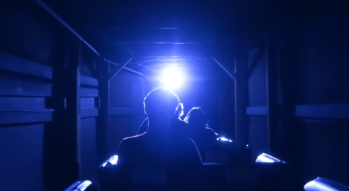 A dark roller coaster tunnel illuminated by a bright blue light, with riders visible in silhouette.