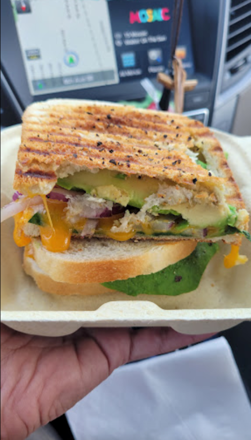 A close-up of a grilled sandwich with layers of vegetables and cheese, held in a hand, inside a car.