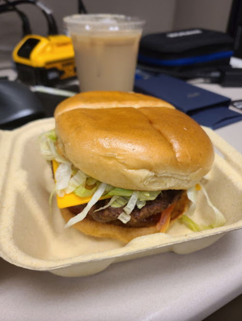 A close-up of a cheeseburger with lettuce in a takeout container, next to a cup of iced coffee on a desk.