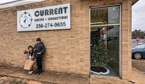 Two children stand outside a shop called "Current Coffee + Collectibles," holding holiday decorations.