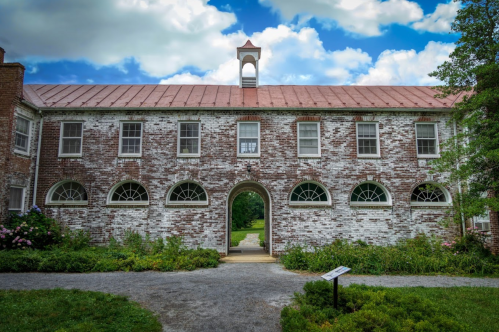 Historic brick building with arched windows and a bell tower, surrounded by greenery and a cloudy blue sky.