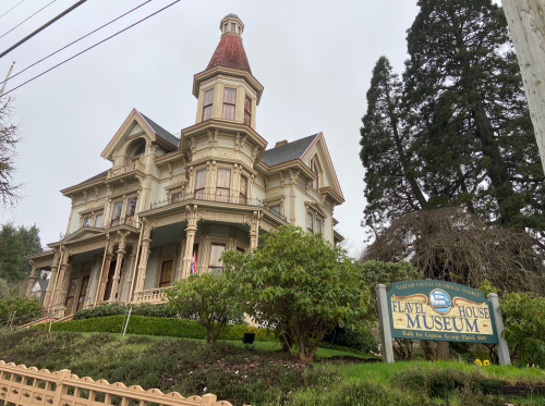 Historic Flavel House Museum, a Victorian-style building with a turret, surrounded by greenery and a sign.