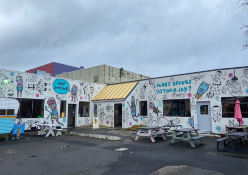 Colorful mural-covered building with picnic tables outside, featuring whimsical characters and the text "What Brings Astoria Joy?"