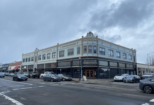 Historic building on a street corner, featuring a mix of shops and cloudy skies above. Cars parked in front.