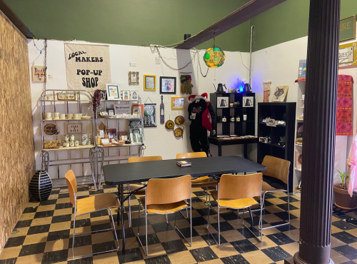 A cozy pop-up shop featuring a table, chairs, shelves with decor, and art on the walls in a vibrant space.