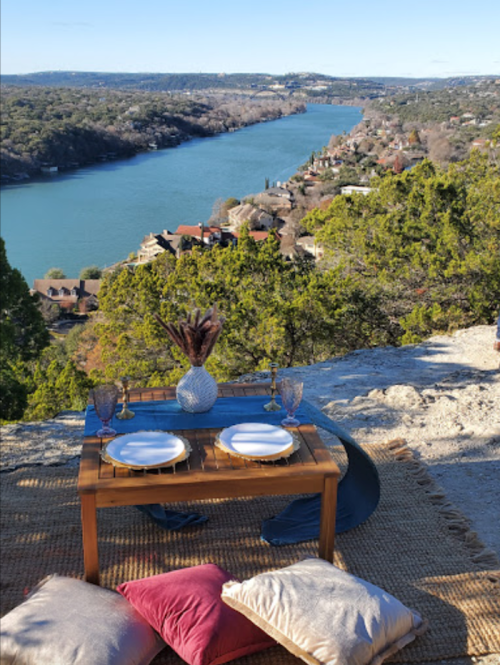 A scenic outdoor dining setup with a table, plates, and cushions overlooking a river and lush landscape.