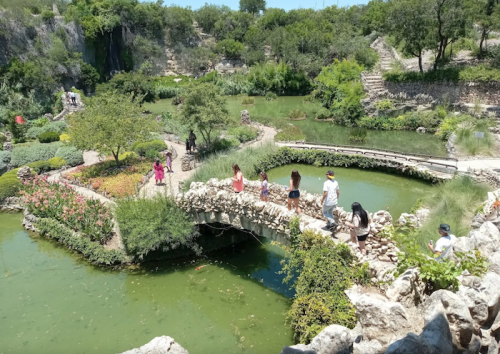 A scenic garden with a stone bridge, lush greenery, and visitors walking along paths by a pond.
