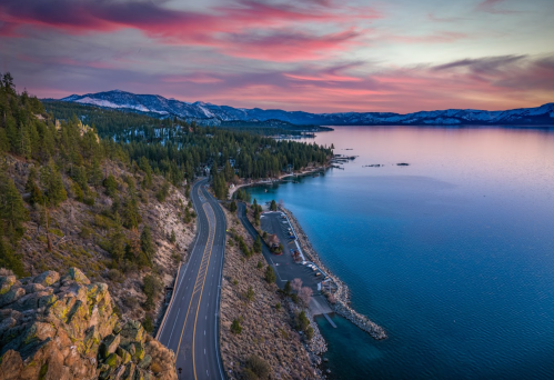 A scenic view of a winding road along a lake at sunset, surrounded by mountains and trees under a colorful sky.