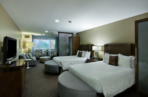 A modern hotel room featuring two beds, a seating area, and a large TV, with soft lighting and neutral decor.