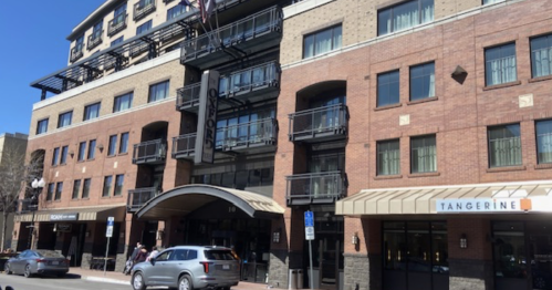 A modern brick building with balconies, featuring shops on the ground floor and a clear blue sky above.