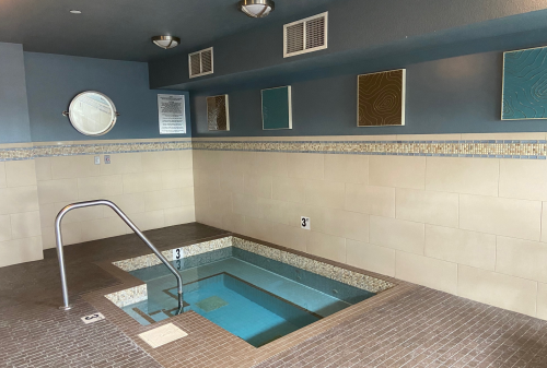 A small indoor spa area featuring a tiled hot tub, wall art, and a mirror, with soft lighting and a clean design.