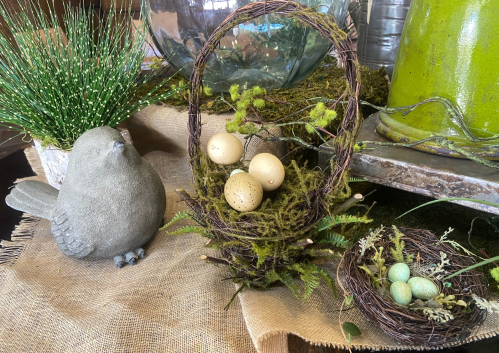 A decorative scene featuring a bird statue, two nests with eggs, and greenery on a rustic surface.