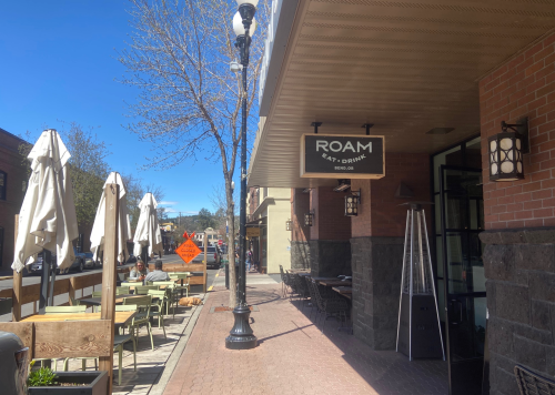 A sunny street scene featuring the ROAM eatery sign, outdoor seating, and trees along a bustling sidewalk.