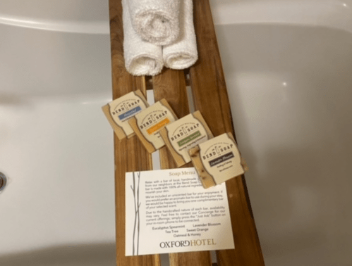A wooden tray with three soap bars and rolled towels, alongside a soap menu from the Oxford Hotel.