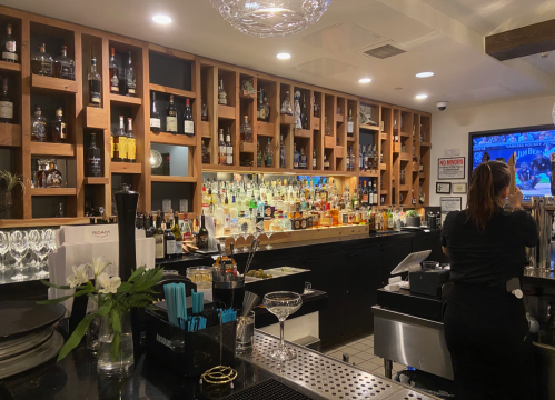 A well-stocked bar with shelves of liquor bottles, a bartender, and a TV displaying a sports game.