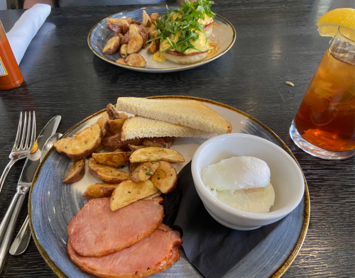 A plate with ham, fried potatoes, two poached eggs, and a side of toast, with a drink and another dish in the background.