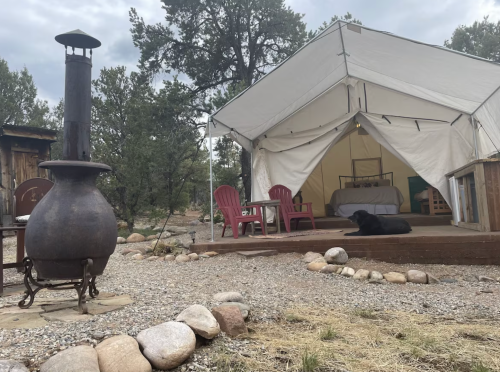 A cozy outdoor tent with a wooden deck, a black dog resting nearby, and a metal chiminea in a rocky landscape.
