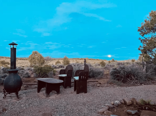 A serene outdoor scene with two chairs and a table near a fire pit, set against a blue sky and distant moonrise.