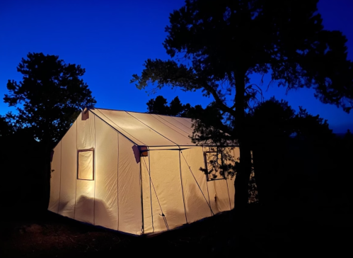 A lit tent stands among trees at dusk, with a deep blue sky in the background.