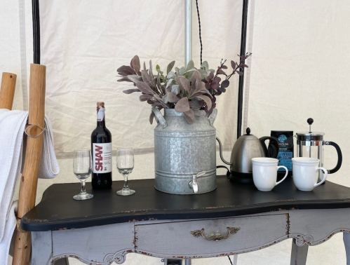 A rustic table with a metal vase of greenery, wine bottle, glasses, a kettle, and coffee mugs in a cozy setting.