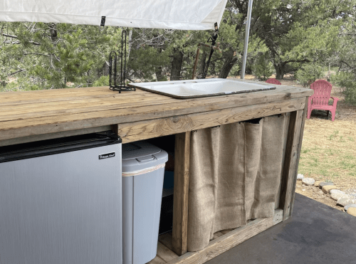 Outdoor kitchen setup featuring a wooden counter, sink, mini fridge, and storage with fabric curtains.