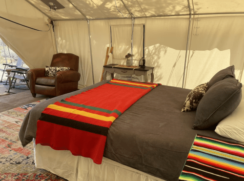 Cozy glamping tent interior with a bed, colorful blanket, armchair, and rustic decor.
