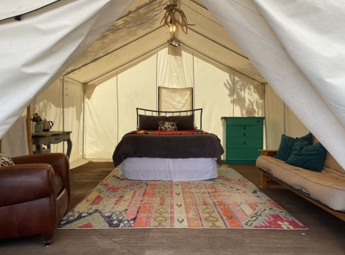 Cozy tent interior featuring a bed, a green dresser, a sofa, and a colorful rug. Natural light filters in.