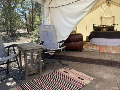 A cozy glamping setup featuring a tent, rocking chairs, a small table, and a bed with decorative pillows.