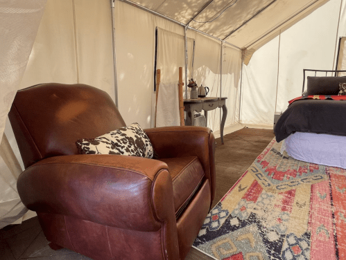 A cozy tent interior featuring a brown leather armchair, a patterned rug, and a bed with colorful bedding.