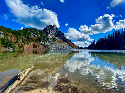 A serene lake reflecting clouds and mountains, surrounded by lush greenery and a clear blue sky.