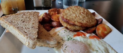 A plate of breakfast featuring eggs, hash browns, roasted potatoes, toast, and a drink in the background.