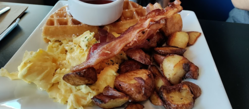 A breakfast plate featuring waffles, scrambled eggs, bacon, and crispy potatoes with a side of syrup.