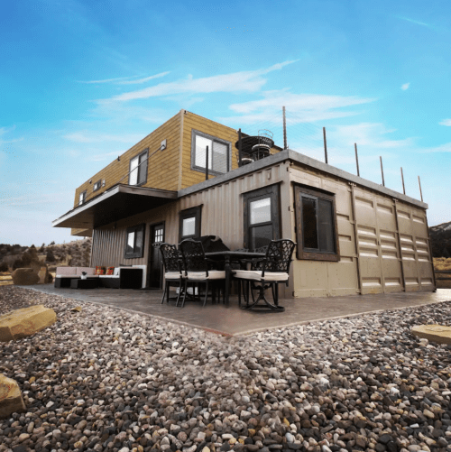 A modern container home with a deck, outdoor seating, and a scenic backdrop of rocks and blue sky.