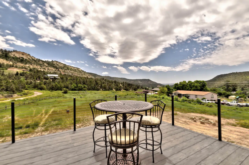 A scenic view from a deck featuring a table and chairs, overlooking green fields and mountains under a cloudy sky.