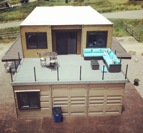 A modern house with a rooftop deck, featuring outdoor seating and a table, surrounded by greenery and a gravel area.