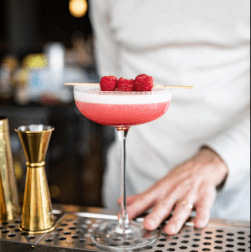 A vibrant pink cocktail garnished with raspberries, served in a coupe glass on a bar counter.