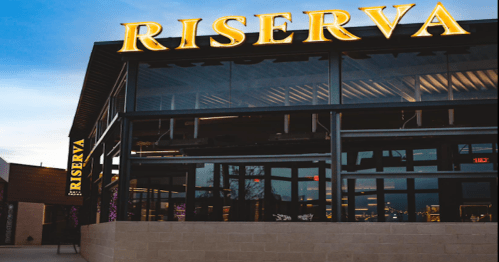 Exterior of a modern restaurant named "Riserva," featuring large illuminated signage against a twilight sky.