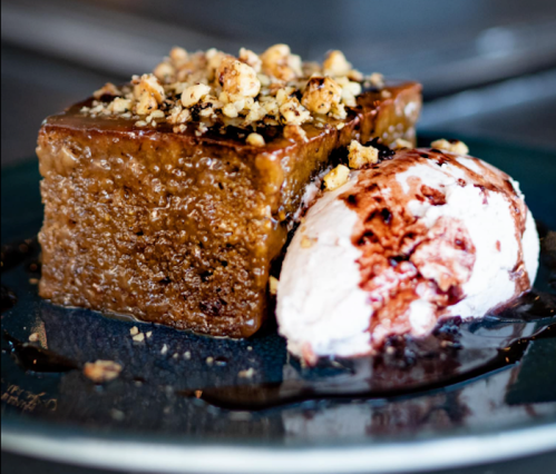 A slice of moist cake topped with nuts, served with a scoop of ice cream drizzled with chocolate sauce.
