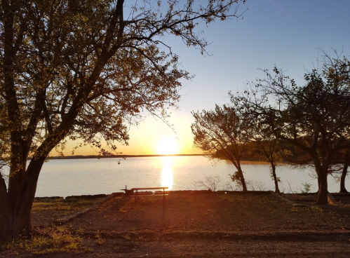 Sunset over a calm lake, framed by trees, casting warm golden light on the water and shoreline.