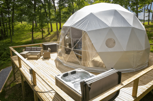 A geodesic dome on a wooden deck, featuring a hot tub and outdoor seating, surrounded by trees.