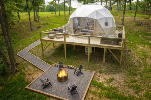 A geodesic dome on a wooden deck surrounded by trees, with a fire pit and seating area in front.