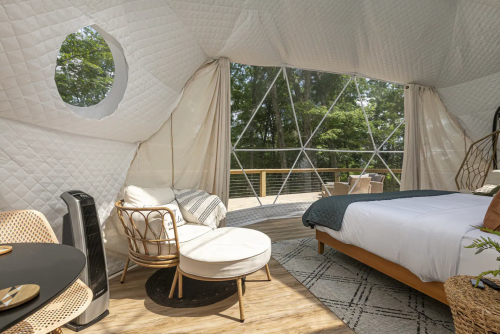 Cozy interior of a geodesic dome with a bed, seating area, and large windows overlooking nature.